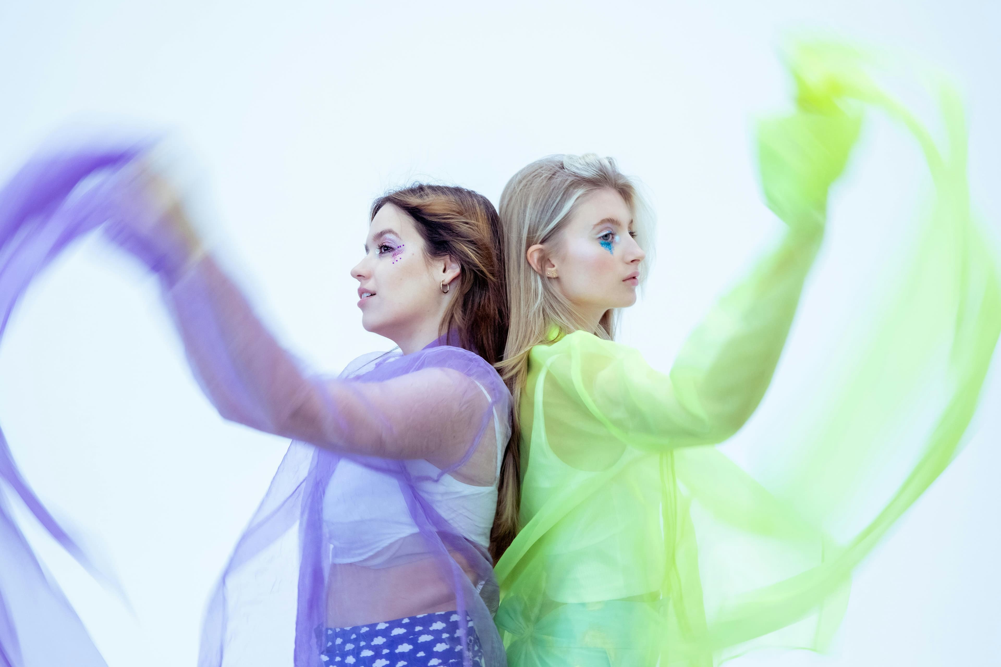 Two women pose back-to-back with vibrant flowing sheer fabrics